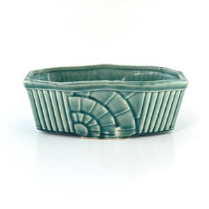May include: A teal green ceramic planter with a geometric design. The planter has a rectangular shape with rounded corners and a raised rim.