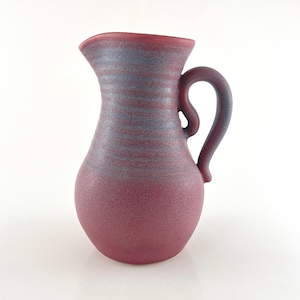 May include: A ceramic pitcher with a rounded body and a curved handle. The surface features a matte finish in a deep magenta hue, with horizontal bands of a muted blue-grey color encircling the upper neck and the handle.