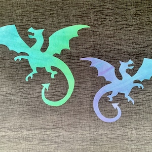 Peut inclure: Deux découpes de dragons dans des tons de bleu et de vert sont affichées sur un fond texturé et sombre. Les dragons ont des ailes déployées et des queues incurvées. Le dragon de gauche est vert et bleu, et celui de droite est bleu et violet.