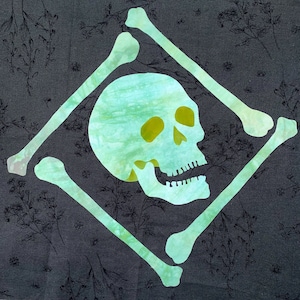 Può includere: Un disegno di teschio e ossa incrociate turchese su sfondo grigio scuro con un motivo floreale. Il teschio è centrato, circondato da quattro ossa a forma di diamante. Il design è probabilmente per Halloween o un tema simile.