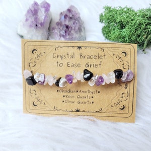 May include: A crystal bracelet featuring black, purple, and white stones. The card reads "Crystal Bracelet to Ease Grief" and lists the stones as "Obsidian, Amethyst, Rose Quartz, Clear Quartz".