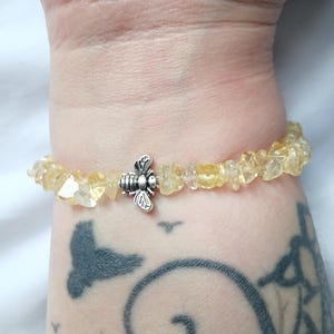 May include: A bracelet made with yellow citrine gemstone chips and a silver-coloured bee charm. The bracelet is strung on an elastic band. The citrine stones have a natural, irregular shape.