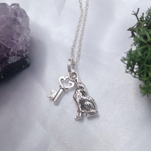 May include: A silver necklace featuring a key charm with a heart-shaped top and a howling wolf charm. The wolf is depicted in a seated pose. The necklace is suspended from a silver chain.