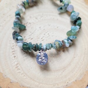 May include: A bracelet made of green and white stone beads. A silver-coloured charm with a rose design is suspended from the centre. The bracelet is composed of small, irregularly shaped stones.