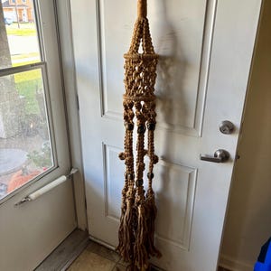 May include: A brown macrame plant hanger with a black metal ring at the top. The plant hanger features a woven design with wooden beads and long, fringed tassels at the bottom. It is hanging against a white door.