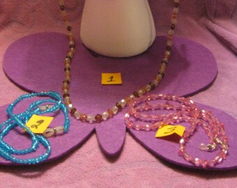 May include: Three beaded eyeglass chains, one brown and white, one pink, and one blue. The chains are displayed on a purple surface with yellow numbered tags.