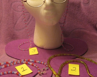May include: A white mannequin head wearing eyeglasses with a beaded eyeglass chain. Three beaded eyeglass chains in red, white, blue, and green are displayed on a purple surface. Each chain has a small yellow tag with a number.