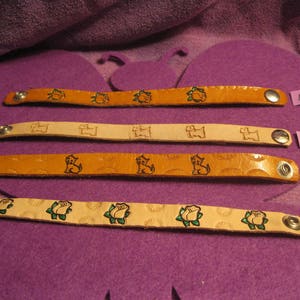 May include: Four leather bracelets with different designs. The bracelets are brown and cream colored with green and brown details. The designs include a dog, a rose, and a turtle. The bracelets are numbered 1 through 4.