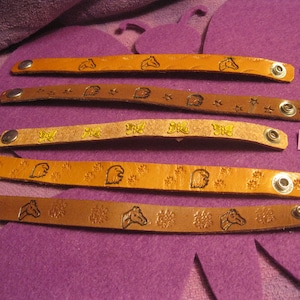 May include: Five leather bracelets with different designs. The bracelets are brown with yellow and black accents. The designs include horses, stars, butterflies, and paw prints. The bracelets are numbered 1 through 5.