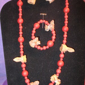 May include: A red beaded necklace with squirrel charms. The necklace is on a black background. The necklace has a matching pair of earrings with squirrel charms.
