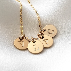 May include: Gold necklace with four small round charms, each with a different letter engraved on it: V, L, P, and C. The necklace is on a white fabric background.