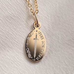 Puede incluir: Un collar con colgante dorado con un colgante ovalado con un diseño de espada y las palabras "YOU DO NOT YIELD". El collar tiene una delicada cadena dorada. La marca "Amoorella Jewelry" está en la parte inferior.