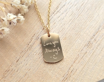 May include: A gold-colored dog tag pendant necklace. The tag is engraved with the words "Love you Always" and a small heart symbol. The necklace is displayed on a wooden surface with dried white flowers.