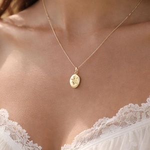 Könnte beinhalten: Eine goldene ovale Anhängerkette mit einem eingravierten Blumenmuster auf der Vorderseite, die an einer zarten Goldkette hängt. Der Anhänger ist mittig auf der Brust platziert und der Text Amoorella Jewelry WWW.AMOORELLA.COM ist unten sichtbar.