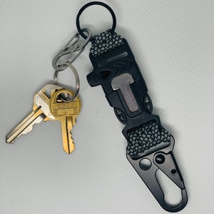 May include: A black key chain with a gray and black paracord strap, a silver clip, and a black carabiner. Two keys are attached to the keychain, one with the number 0950 on it.
