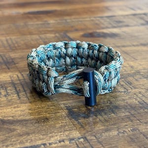 May include: A braided paracord bracelet in shades of green, blue, and tan. The bracelet features a black metal clasp. The bracelet is approximately 0.75 inches wide.