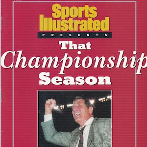 May include: Sports Illustrated magazine cover featuring a red background with the text "Sports Illustrated Presents That Championship Season". A man in a plaid jacket and tie is raising his fist in the air, with a football helmet in the foreground.