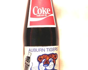 May include: A glass bottle of Coke with a red and white label featuring the text Coke. Below, a white label displays an illustration of an orange and black tiger holding a bottle, with the text AUBURN TIGERS, 1983 SEC CHAMPIONS, and 1984 SUGAR BOWL CHAMPS.