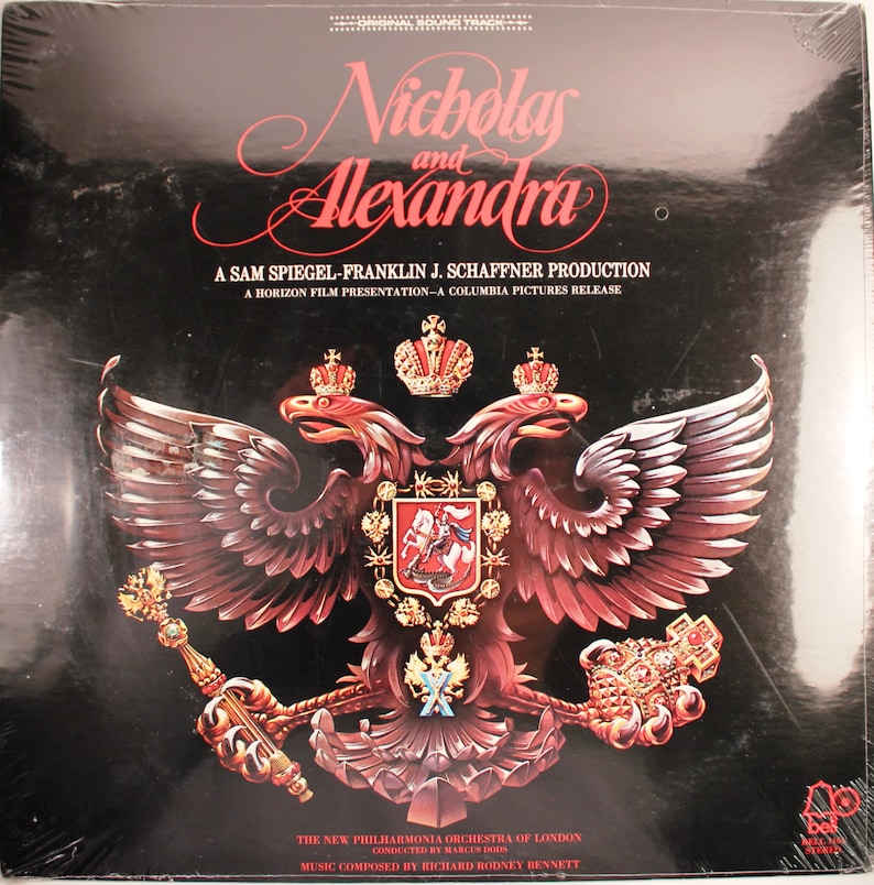 May include: A black vinyl record cover with a gold and red image of a double-headed eagle with a crown on top. The text "Nicholas and Alexandra" is written in a cursive font at the top of the cover. The text "A Sam Spiegel-Franklin J. Schaffner Production" is written in a smaller font below the image. The text "A Horizon Film Presentation-A Columbia Pictures Release" is written in an even smaller font below the production text. The text "The New Philharmonia Orchestra of London" is written in a smaller font at the bottom of the cover. The text "Conducted by Marces Dods" is written in an even smaller font below the orchestra text. The text "Music Composed by Richard Rodney Bennett" is written in an even smaller font below the conductor text.