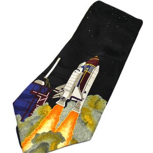 May include: Black necktie featuring a space shuttle design. The tie has a black background with a starry night sky. The shuttle is white, gray, and red, with orange flames. A building with blue and red accents is also visible.