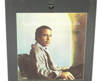May include: A vintage 8-track tape cartridge featuring Paul Simon's "Greatest Hits, Etc." album. The cartridge is dark gray, with a color image of Paul Simon on the label. The text "Paul Simon - Greatest Hits, Etc." is printed on the label.
