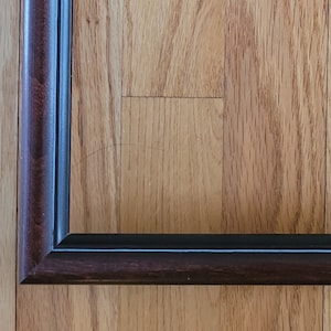 May include: Rectangular picture frame with a dark brown, almost black, finish. The frame has a simple, classic design with a narrow profile. The frame is empty, ready to display a photograph or artwork. The frame is sitting on a wooden surface.