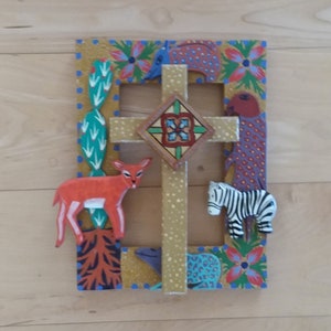 May include: A decorative wooden cross with a gold-colored frame, featuring a deer and zebra. The frame is adorned with colorful floral and animal designs. The cross has a central diamond-shaped emblem.