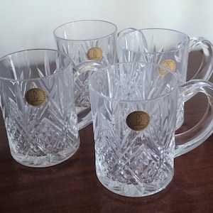 May include: Four clear crystal mugs with a diamond-cut pattern and curved handles. Each mug has a gold-colored sticker with a letter "P" on it. The mugs are arranged on a dark brown wooden surface.
