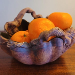 May include: A decorative, handcrafted bowl filled with bright orange tangerines. The bowl has a unique, organic shape with a dark purple and brown color scheme. The handle is a twisted, vine-like design, adding to its rustic charm. The bowl sits on a wooden surface.