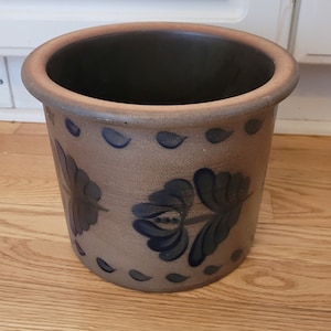 May include: A cylindrical, tan-colored ceramic planter with a dark interior. The planter features blue floral and leaf designs around the exterior. The rim is a lighter tan color.