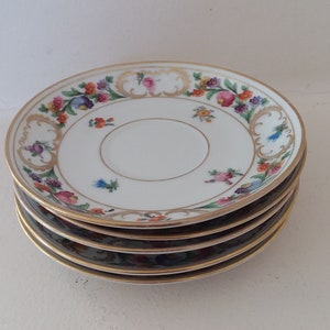 May include: A stack of five white porcelain saucers. Each saucer features a gold rim and a floral design with pink, orange, blue, and purple flowers. The saucers are stacked on top of each other.