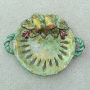 May include: A hand-painted, ceramic decorative bowl with a green and yellow color scheme. The bowl has a round shape with cut-out designs and two rope-like handles. The top is decorated with leaves and yellow and brown floral accents.