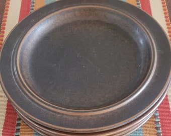 May include: A stack of four dark brown ceramic plates with a raised rim. The plates are on a colorful striped tablecloth with red, tan, teal, and brown stripes. The plates have a matte finish and a simple, classic design.