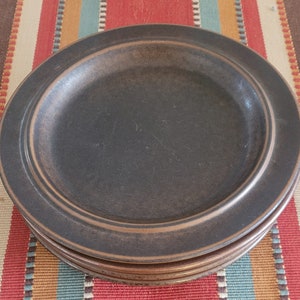 May include: A stack of four dark brown ceramic plates with a raised rim. The plates are on a colorful striped tablecloth with red, tan, teal, and brown stripes. The plates have a matte finish and a simple, classic design.