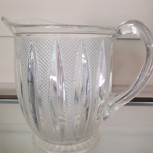 May include: Clear glass pitcher with a handle and a spout. The pitcher has a textured diamond pattern on the body with vertical lines. The handle is curved and attached to the side of the pitcher. The pitcher sits on a small, round base.