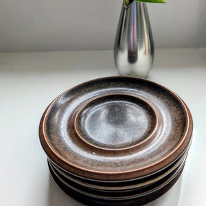 May include: A stack of four brown and copper-toned saucers with a raised center. A silver vase holds a pink flower with a dark center and green leaves. The items are on a white surface.