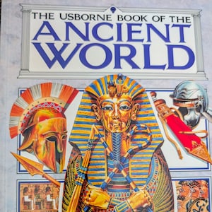 May include: A hardcover book titled "The Usborne Book of the Ancient World." The cover features illustrations of ancient Egyptian pharaohs, Greek helmets, and other artifacts. The book has a light blue background with the title in large blue letters.
