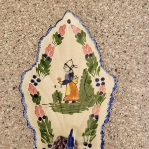 May include: A hand-painted ceramic wall pocket in a leaf shape. The off-white pocket features a blue scalloped edge, a painted scene of a person in traditional clothing, and floral and berry accents. A blue hook is at the bottom.