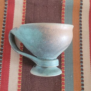 May include: A handmade ceramic cup with a handle and a pedestal base. The cup is glazed in shades of blue and tan, with a speckled texture. The cup is sitting on a striped fabric.