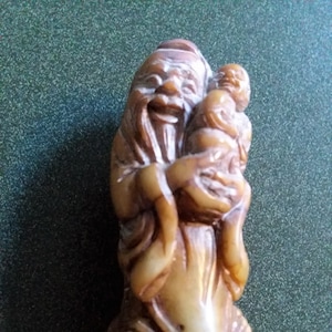 May include: A carved figurine depicting an elderly man holding a small child. The sculpture is a light brown color with detailed facial features and clothing. The base of the figurine has etched designs. The background is a dark green.