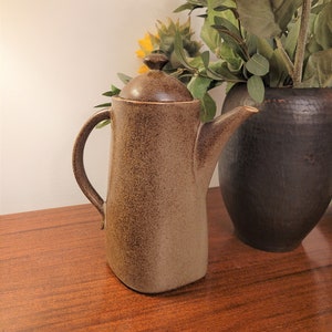 May include: A brown ceramic coffee pot with a lid, handle, and spout. The pot has a speckled texture and sits on a wooden surface. A vase with sunflowers is in the background.