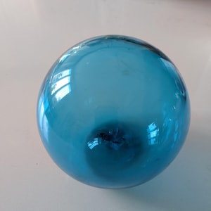 May include: A round, transparent, light blue glass ball. The sphere reflects light, creating bright highlights on its surface. The glass has a slight imperfection near the bottom. The object is likely a decorative item.