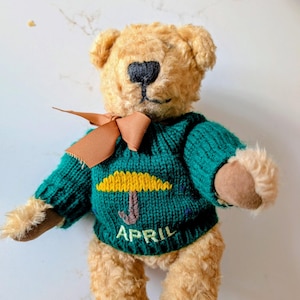 May include: A plush teddy bear with tan fur, wearing a green sweater with a yellow umbrella design and the word "APRIL". The bear has a brown bow tie and a black stitched nose.