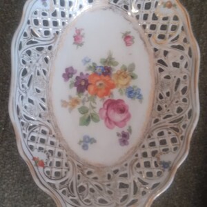May include: Oval-shaped, white porcelain serving dish with an openwork design and gold trim. The center features a floral arrangement with pink, orange, purple, and blue flowers. A decorative piece for serving or display.
