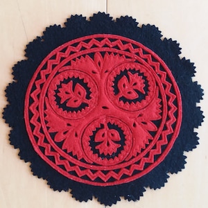 May include: A round, felt coaster with a black scalloped edge. The center features a red, intricate floral design. The coaster is likely handmade and adds a decorative touch to any table setting. The coaster is approximately 6 inches in diameter.