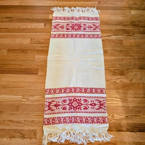 May include: Cream-colored rectangular textile with red embroidered floral patterns and fringe trim. The embroidery features geometric and floral designs. The textile is likely a decorative table runner or wall hanging.