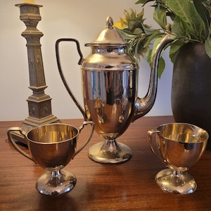 May include: A silver tea set with a tall coffee pot, two handled cups, and a creamer. The set is on a dark wooden surface. A tall, ornate candlestick and a vase of greenery are in the background.