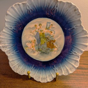 May include: A decorative ceramic plate with a ruffled edge. The plate features a central image of figures in classical attire, surrounded by a gradient of blue and white, with gold accents. The plate is displayed on a gold-colored stand.