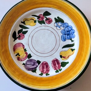 May include: A decorative ceramic plate with a white center and a yellow and green border. The plate features hand-painted fruit designs, including grapes, plums, and pears. The plate is a round shape.