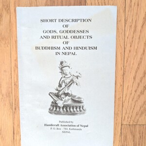 May include: A white pamphlet with the title "SHORT DESCRIPTION OF GODS, GODDESSES AND RITUAL OBJECTS OF BUDDHISM AND HINDUISM IN NEPAL." The pamphlet features an illustration of a seated deity and is published by the Handicraft Association of Nepal.